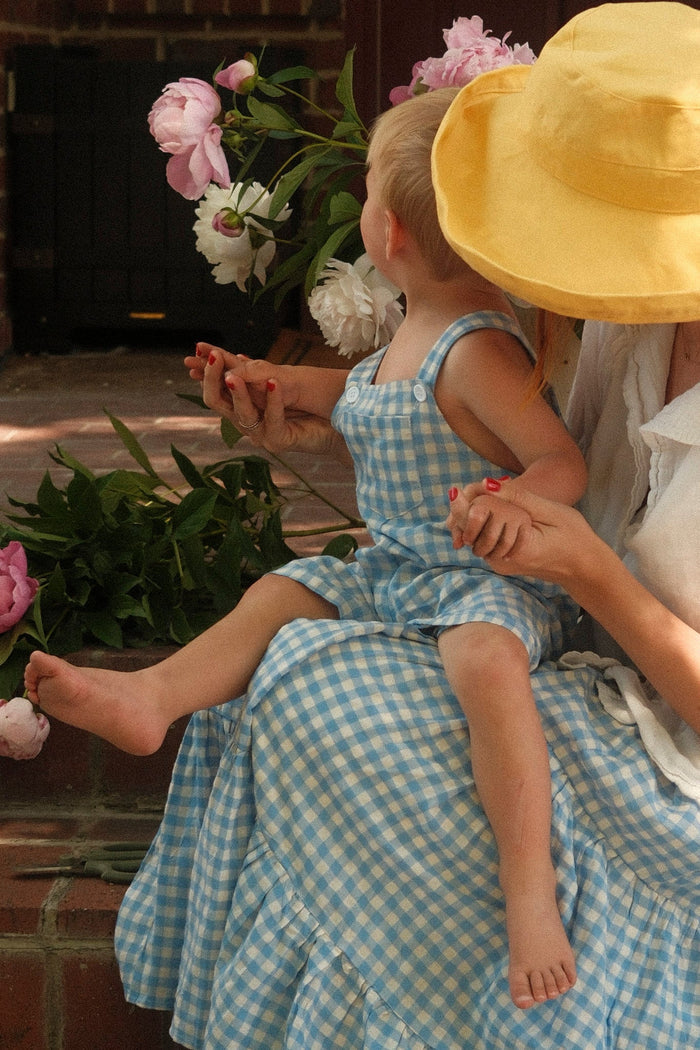 A woman in a yellow hat holds a small child on her lap, who is dressed in the Ivy City Co Baby Alex Overalls in Blue Gingham. They sit together on brick steps surrounded by pink and white flowers, both facing away from the camera in their charming spring outfits.