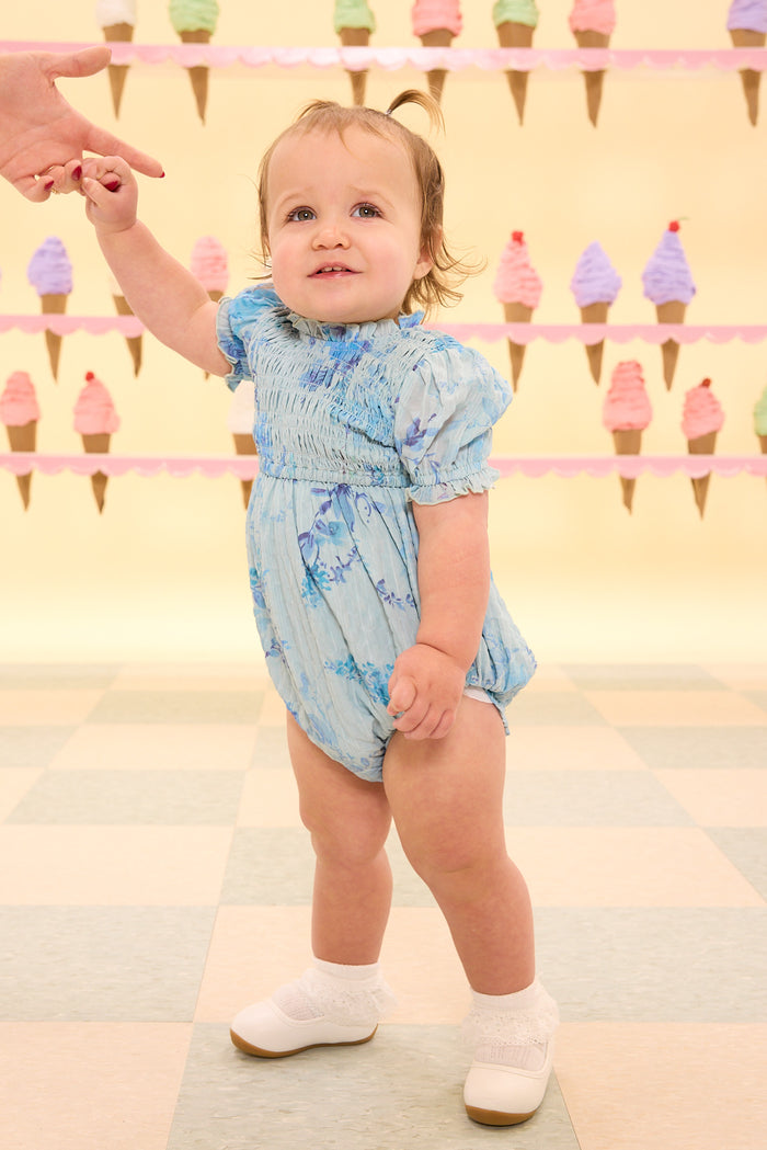 Baby in a blue floral romper standing on a checkered floor with ice cream cone decorations in the background.