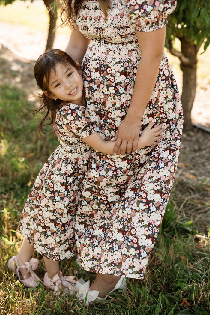 A young girl hugs a woman’s waist outdoors, both wearing matching Ivy City Co Sunday Best Dresses in the Midnight Garden print with smocked bodices and pink shoes. They stand on grass surrounded by trees and sunlight, their maternity-friendly outfits perfect for special moments together.