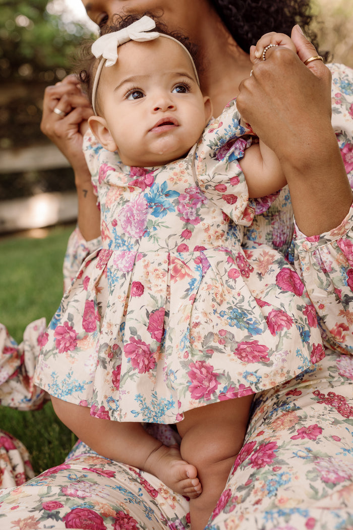 A baby dressed in the Ivy City Co Baby Adelpha Dress Set in Autumn Amity Floral—with its charming smocked cotton design and matching floral headband—is held by an adult also wearing a coordinating floral outfit. Together, they sit outdoors on the grass, creating a sweet, coordinated look that’s perfect for any occasion.
