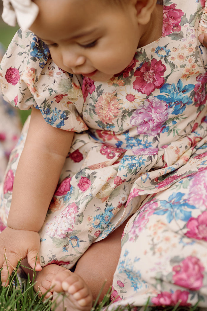 A baby wearing the Ivy City Co Baby Adelpha Dress Set in Autumn Amity Floral sits on the grass, reaching down to touch the blades. The close-up highlights her bare feet and the vibrant flower pattern of her smocked cotton dress.
