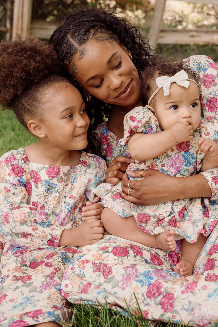 A Black mother and her two daughters sit on the grass. The mother wears a floral dress and holds her baby, who is dressed in the Ivy City Co Baby Adelpha Dress Set in Autumn Amity Floral—a smocked cotton dress paired with a white headband. The older daughter, also wearing a matching Baby Adelpha Dress, gazes lovingly at her baby sister.