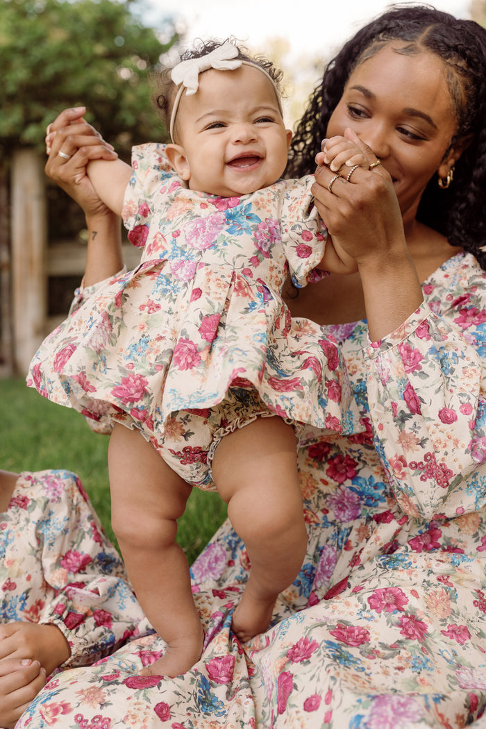 A smiling baby wears the Ivy City Co Baby Adelpha Dress Set in Autumn Amity Floral, complete with a bow, while standing supported by an adult woman who is also dressed in a matching floral outfit. They are outdoors on a grassy area.
