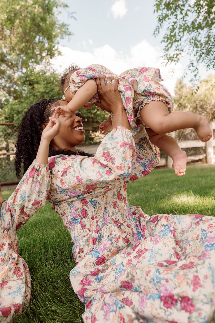A woman in a floral smocked cotton dress sits on the grass, smiling as she lifts a young child dressed in the Ivy City Co Baby Adelpha Dress Set in Autumn Amity Floral above her. Trees and a fence are visible in the background on this sunny day.