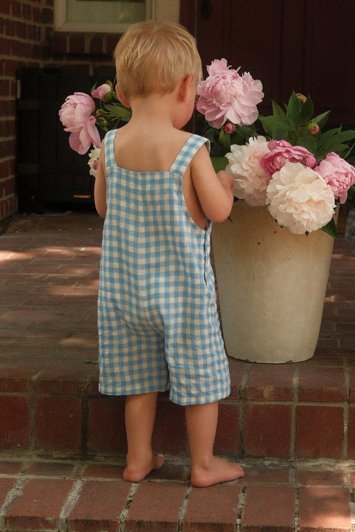 A young child wears the Ivy City Co Baby Alex Overalls in Blue Gingham, standing barefoot on brick steps and admiring pink and white peonies in a large pot—creating the perfect spring outfit.