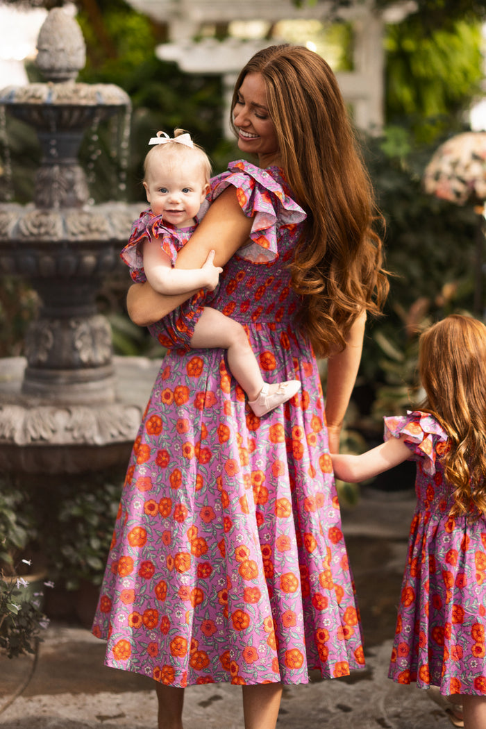 A woman with long brown hair holds a smiling baby girl wearing the Ivy City Co Baby Hattie Romper in Thistle Flower Poppy Woodblock; both match in orange floral dresses while another young girl in a similar dress stands by a stone fountain and greenery.