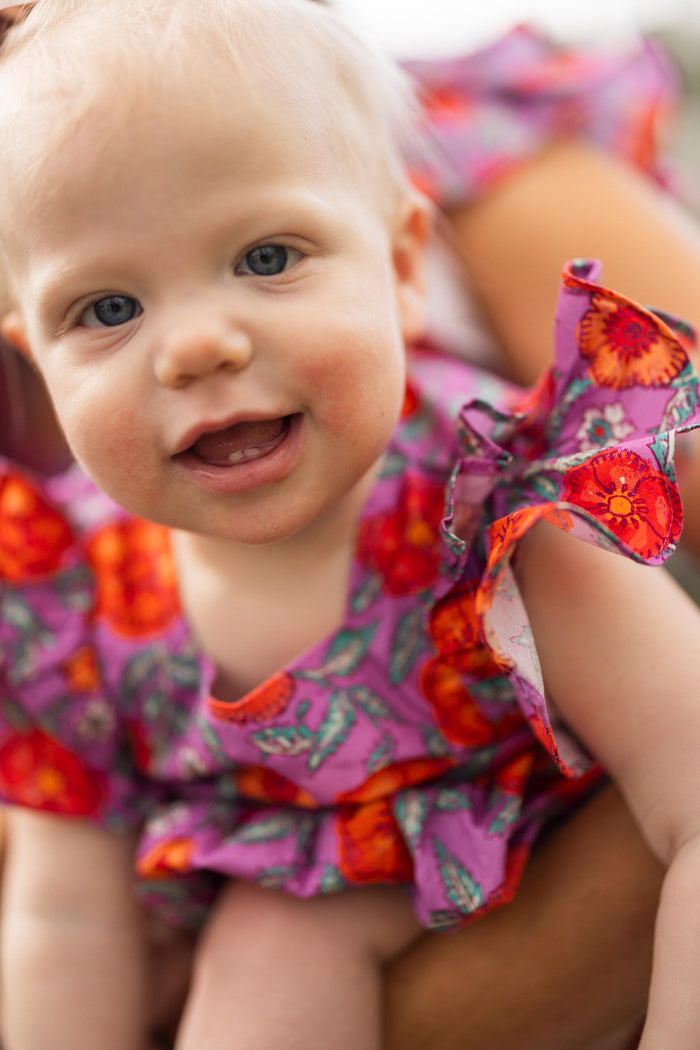 A smiling baby with blue eyes and light hair looks up at the camera, wearing the Ivy City Co Baby Hattie Romper in Thistle Flower Poppy Woodblock—a vibrant purple floral outfit with a smocked bodice and bold orange-red patterns.