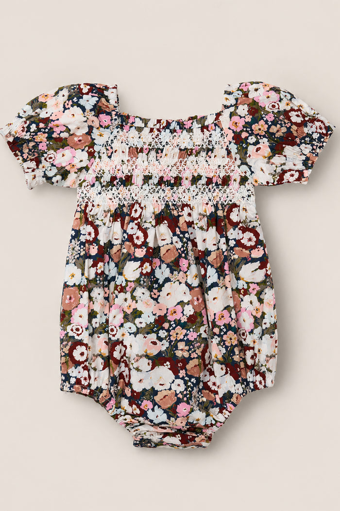 The Ivy City Co Baby Sunday Best Romper in Midnight Garden is a short-sleeved cotton-blend romper with elastic at the legs and shoulders, showcasing a vibrant floral print in shades of pink, white, brown, and green, and featuring a smocked bodice detail across the chest.