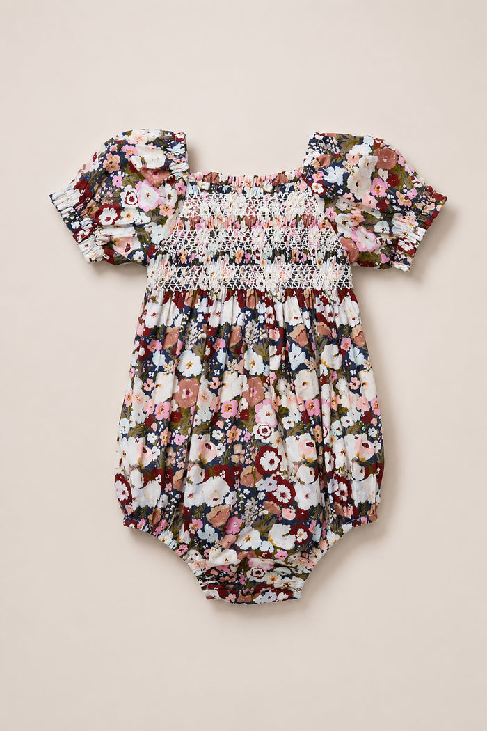 The Ivy City Co Baby Sunday Best Romper in Midnight Garden, featuring puffed short sleeves, a smocked bodice, and a vibrant floral print on a cream cotton-blend fabric, is displayed laid flat on a neutral surface.