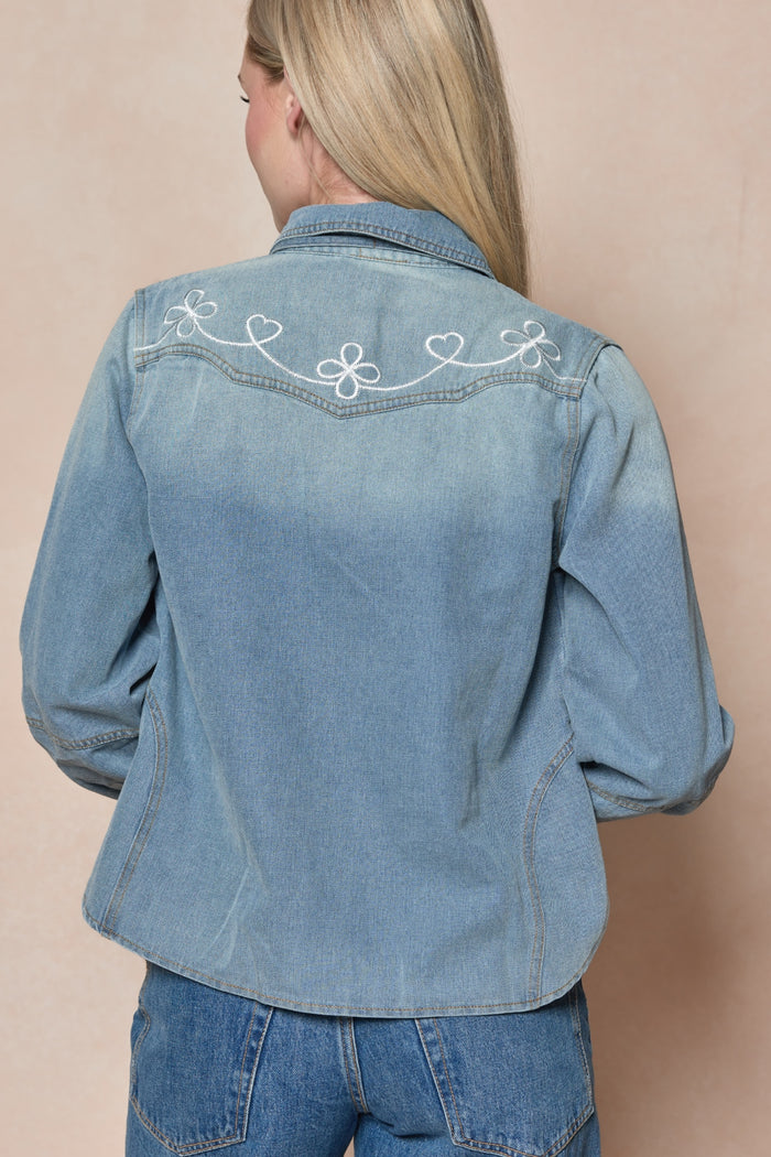 A woman with long blonde hair is seen from the back, wearing the Ivy City Co Denver Top in Lightwash Denim adorned with white embroidered hearts and swirls, paired with matching blue jeans against a plain beige background.