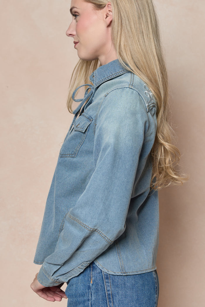 A woman with long blonde hair stands sideways against a light beige background, wearing the Denver Top in Lightwash Denim by Ivy City Co and blue jeans.