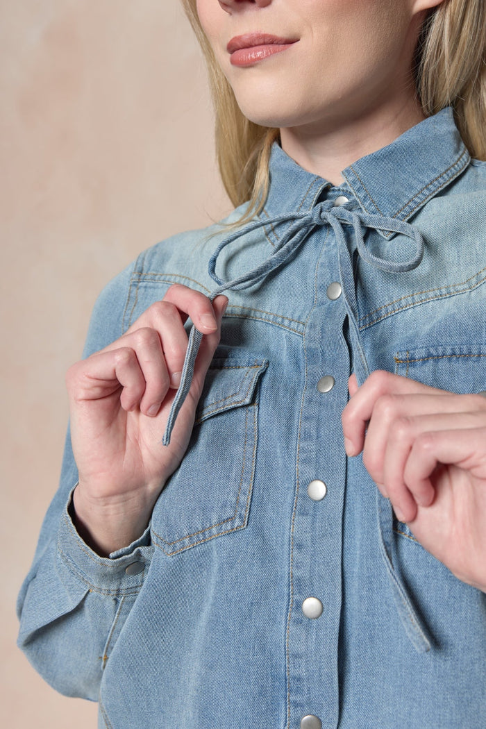 A woman, shown from the lower face to upper torso, adjusts the self-tie bow collar at the neckline of Ivy City Co’s Denver Top in Lightwash Denim—a western-inspired style featuring pearl snap buttons.