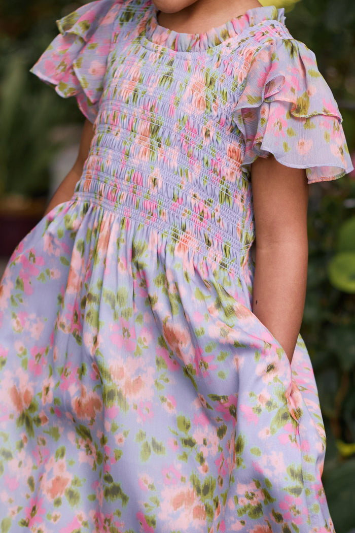 A child in the Ivy City Co Mini Lottie Dress in Vibrant English Garden—a pastel floral midi dress with ruffled sleeves, smocked bodice, and pockets in pink, green, and lilac—stands outdoors.