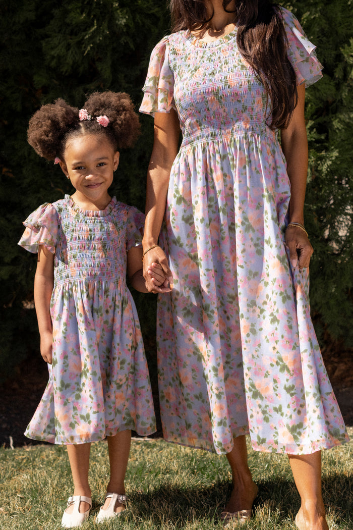 A woman and a young girl hold hands, wearing matching Ivy City Co dresses. The girl wears the Mini Lottie Dress in Vibrant English Garden, featuring short ruffled sleeves. They stand on grass with green trees in the background.