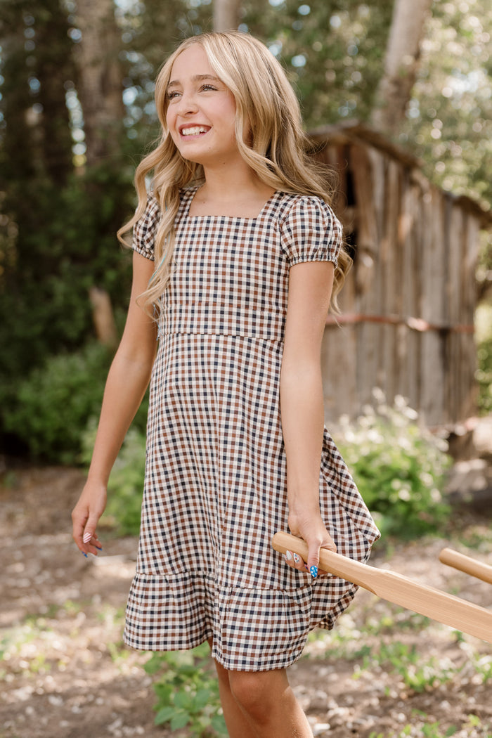 A young girl with long blonde hair stands outdoors, smiling and holding a wooden handle, wearing the Ivy City Co Teen Connie Short Sleeve Dress in Chestnut Gingham, with trees and a rustic wooden shed in the background.