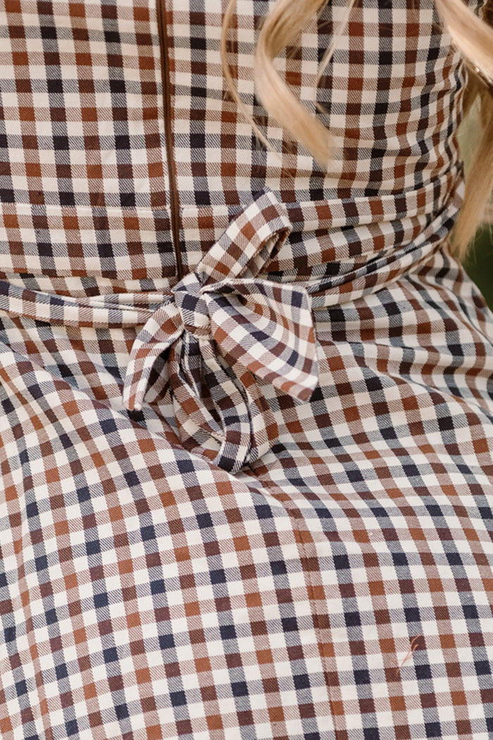 Close-up of the Ivy City Co Teen Connie Short Sleeve Dress in Chestnut Gingham, featuring a brown, blue, and white plaid pattern and a tied bow at the waist. A few strands of blonde hair are visible on the right side of the image.