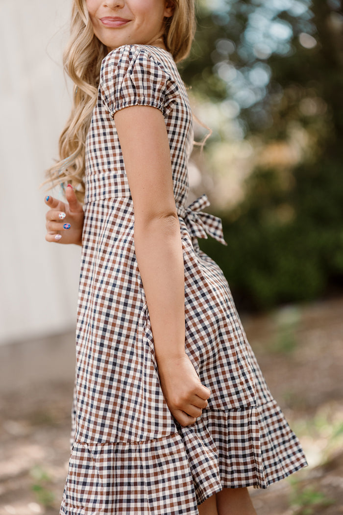 A girl with long blonde hair wears the Ivy City Co Teen Connie Short Sleeve Dress in Chestnut Gingham, its classic plaid pattern and charming back bow standing out. She stands outside amidst greenery, the soft cotton fabric and short sleeves making it ideal for a sunny day, with the background beautifully blurred.