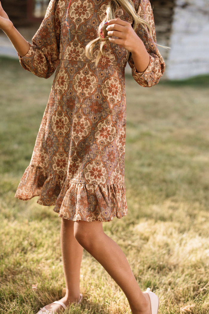 A girl walks barefoot on grass, holding part of her long, wavy hair, wearing the Ivy City Co Teen Draper Dress in Golden Hour Tesserae with balloon sleeves and a floral pattern. Her face is not visible in the image.