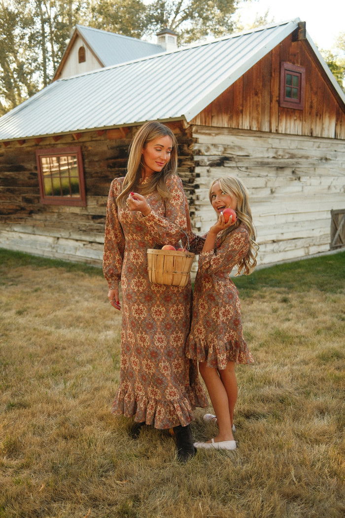 A woman and a young girl, both dressed in matching Ivy City Co Teen Draper Dresses in Golden Hour Tesserae with charming balloon sleeves, hold a basket of peaches as they stand together in a grassy field before a rustic, white-washed log cabin topped with a metal roof. The girl playfully brings an apple to her lips—their coordinated outfits perfectly capturing the picturesque spirit of country autumn days and highlighting the timeless appeal of this fall dress.
