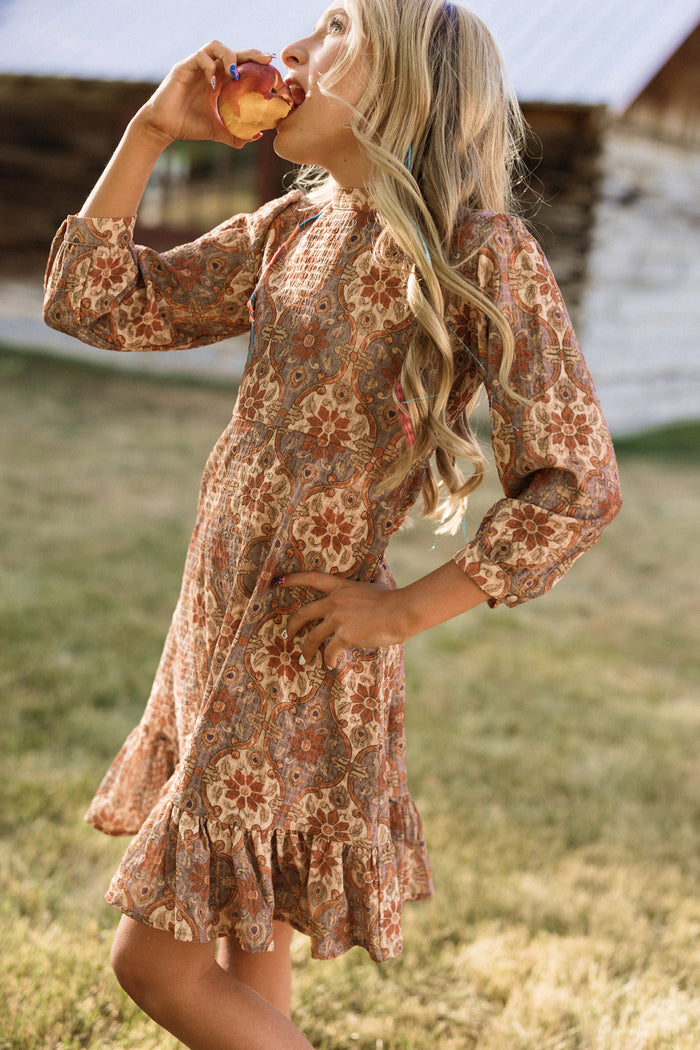 A girl wearing the Ivy City Co Teen Draper Dress in Golden Hour Tesserae, featuring balloon sleeves and a vibrant pattern, stands on grass with a rustic wooden building behind her, holding and biting into a peach.