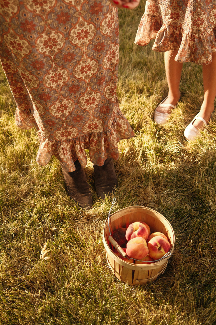 Two people in patterned dresses—one wearing Ivy City Co’s Teen Draper Dress in Golden Hour Tesserae, featuring charming balloon sleeves—stand on grass near a basket brimming with peaches. One person wears boots while the other opts for light shoes. Warm light bathes the scene, evoking a late afternoon or early evening atmosphere and perfectly capturing the essence of an autumn dress moment.